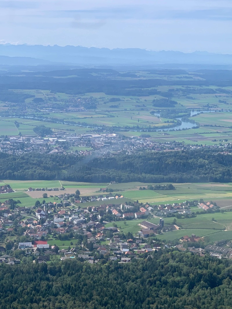 a landscape. houses dot the landscape. there is a large forest and a twisting river and patches of farmers fields